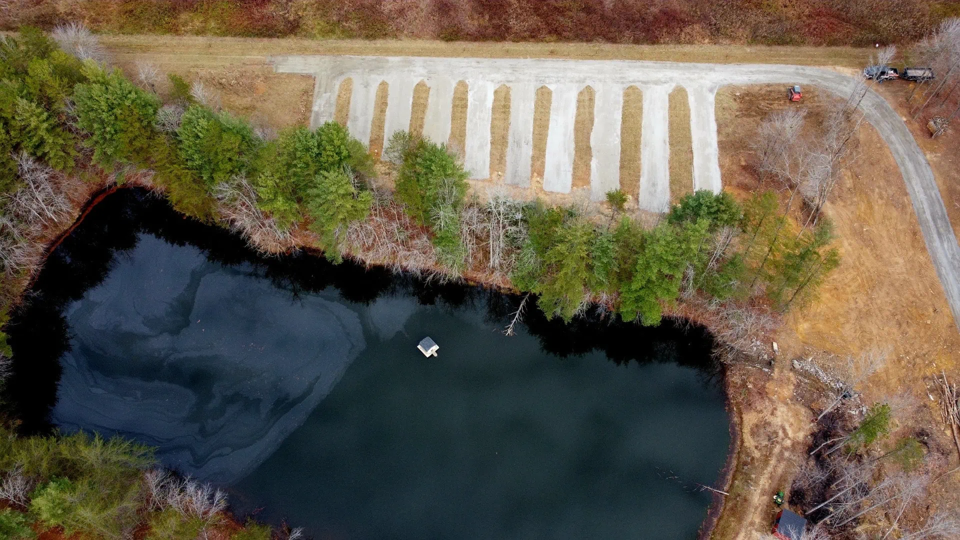Aerial view of a small lake surrounded by trees, with a concrete parking area and a parked vehicle at the edge of a road.