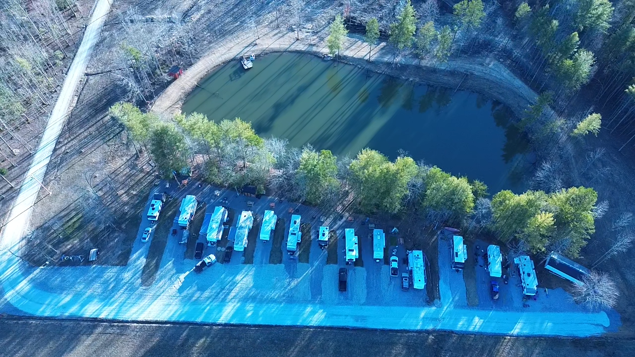 Aerial view of RVs parked in a row near a small pond surrounded by trees, with a road running alongside the parking area.