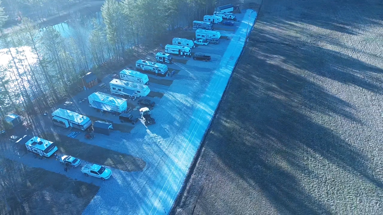 Aerial view of RVs lined up on a gravel parking area adjacent to a wooded area, with a field nearby.